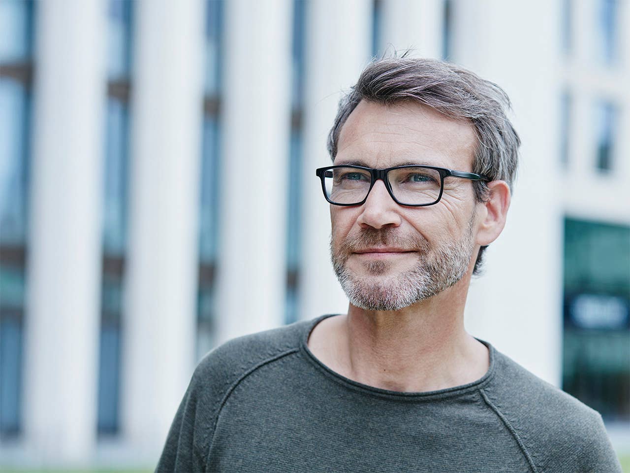 Middle aged white man with glasses and a grey beard stands outdoors in front of an office building