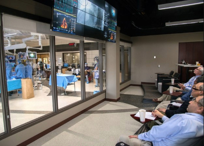 Fellows and other medical staff are watching a procedure in an operating room through a glass window and a tv screen