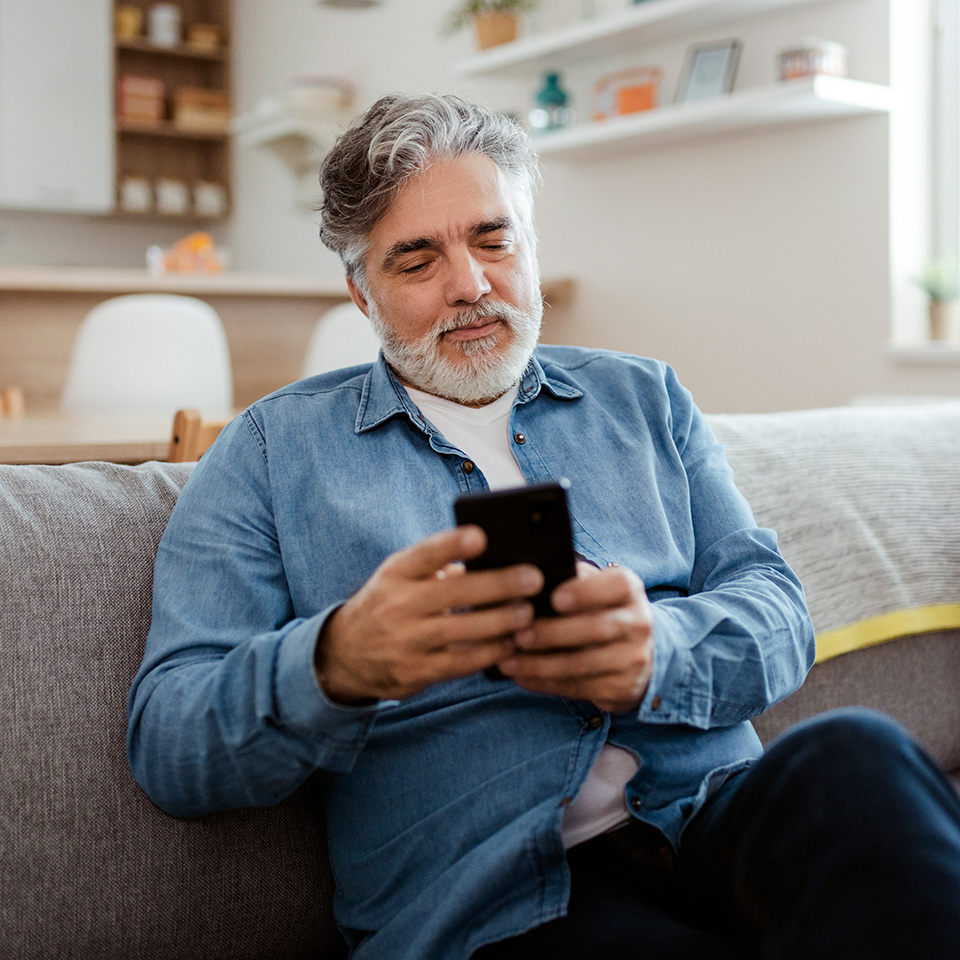 Man with grey hair and grey beard sits on couch looking at his phone