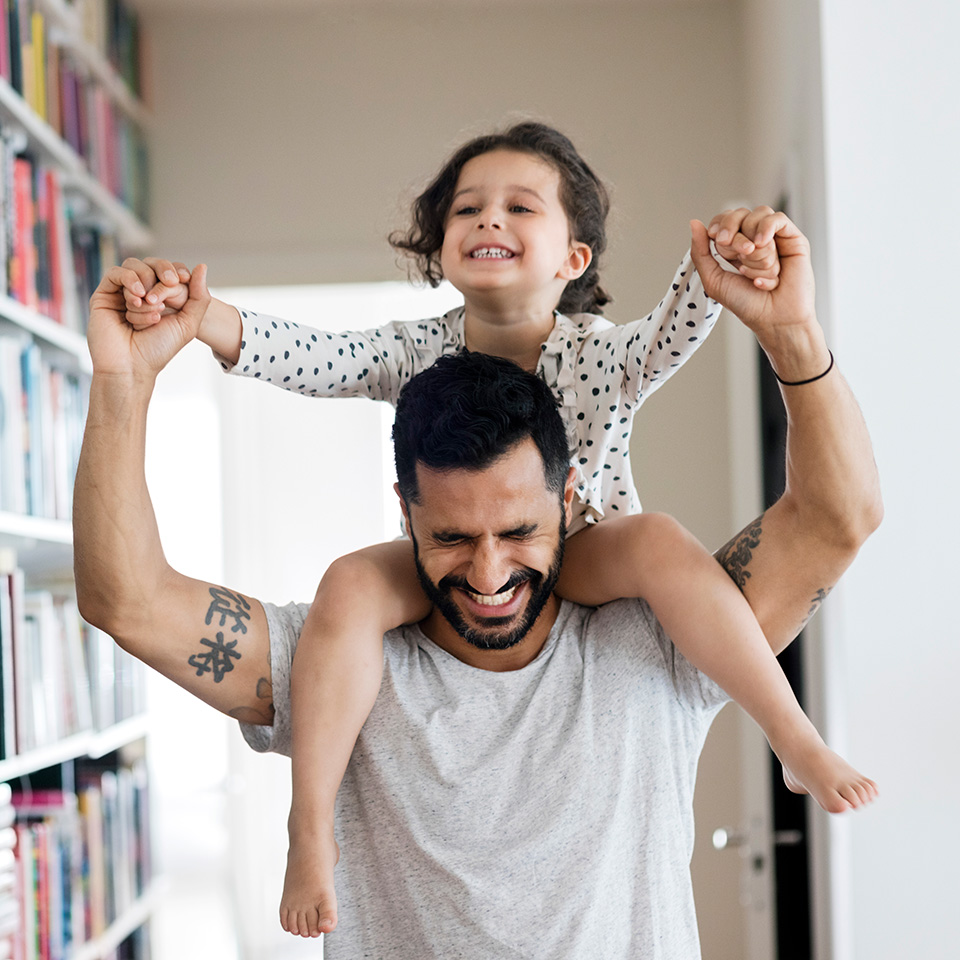 Smiling father with black hair holds young daughter's hands as she sits on his shoulders