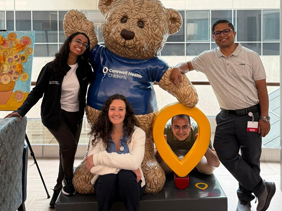 Residents posing with a Corewell Health Children's teddy bear and location marker display.