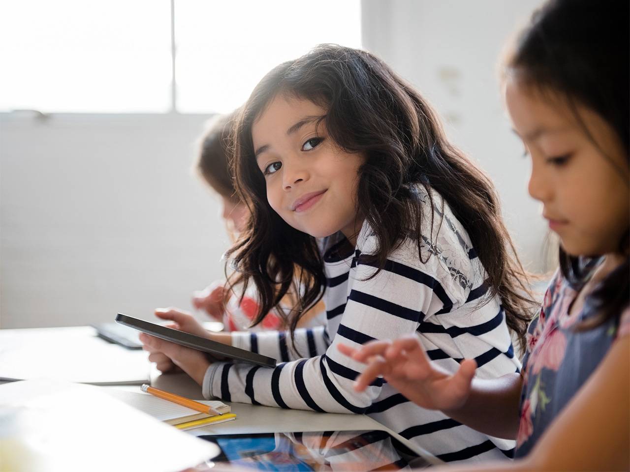 Smiling girl with long brown hair holds a tablet device while in a classroom