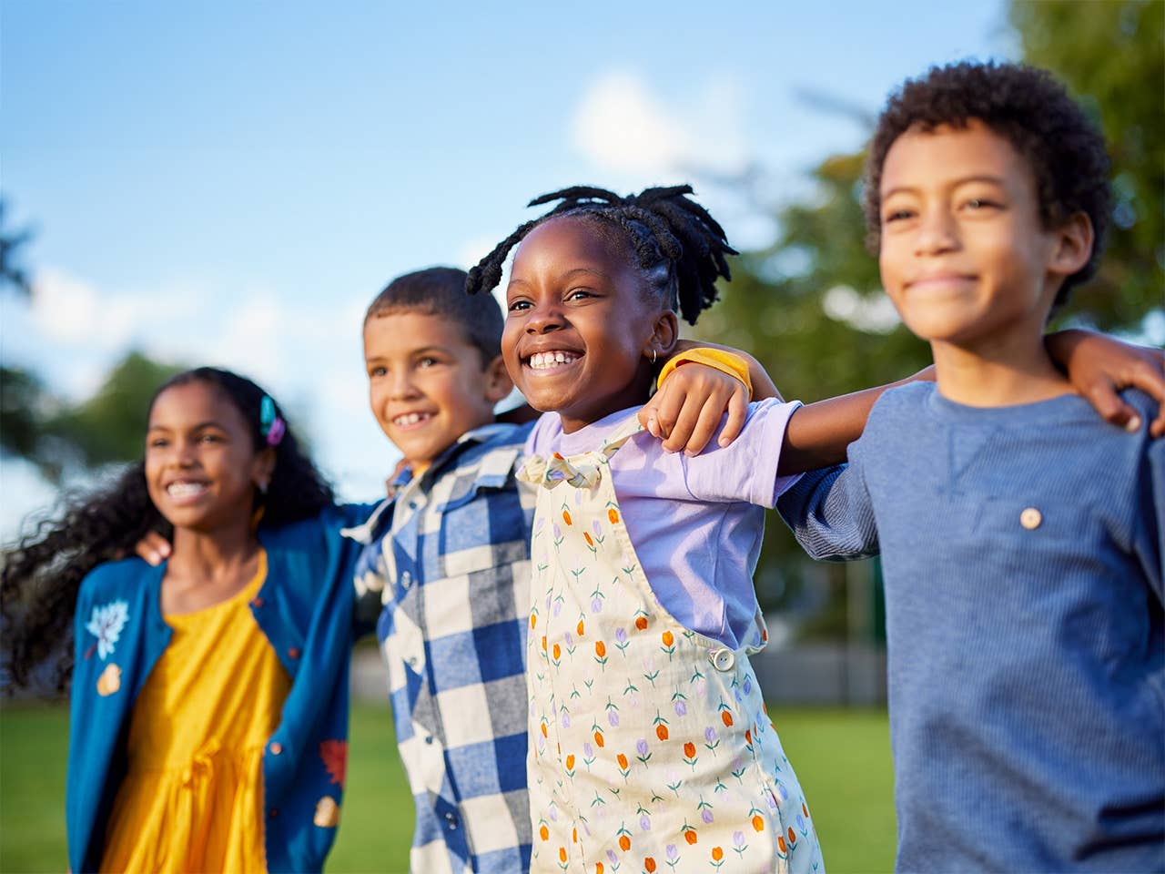 Group of four smiling boys and girls link arms across one another's shoulders outdoors in the sunshine