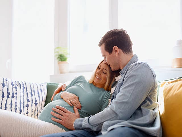 Young man hugs his pregnant wife and places a hand on her belly as they sit on a couch