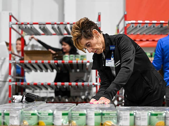 A woman stands in a warehouse surrounded by stacked cans and boxes, organizing supplies for distribution.