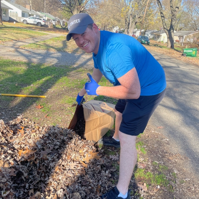 man in a hate outside raking leaves into a paper bag