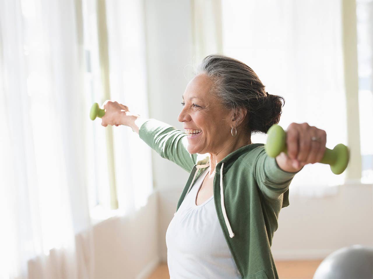 Smiling woman wearing a white shirt and green jacket holds dumbbells in her hand as she stretches her arms out to her sides