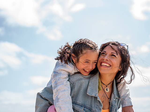 Smiling woman wearing a denim jacket carries her young daughter on her back outdoors in the sunshine