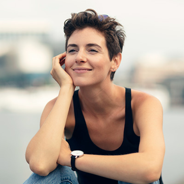 Young woman with short brown hair sits, smiling, with her hand on her cheek