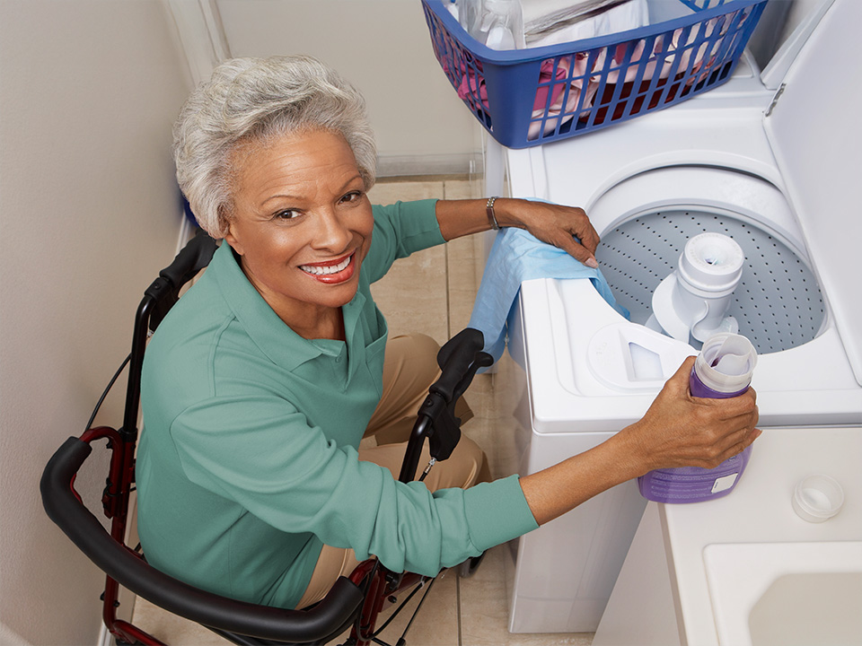 Person seated in a mobility aid adding liquid detergent to a top-loading washing machine in a laundry area, with a laundry basket on top of the washer and cleaning tools visible nearby.