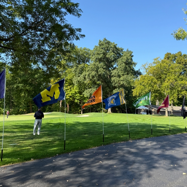 A variety of Michigan school flags on display in the green grass at the golf event