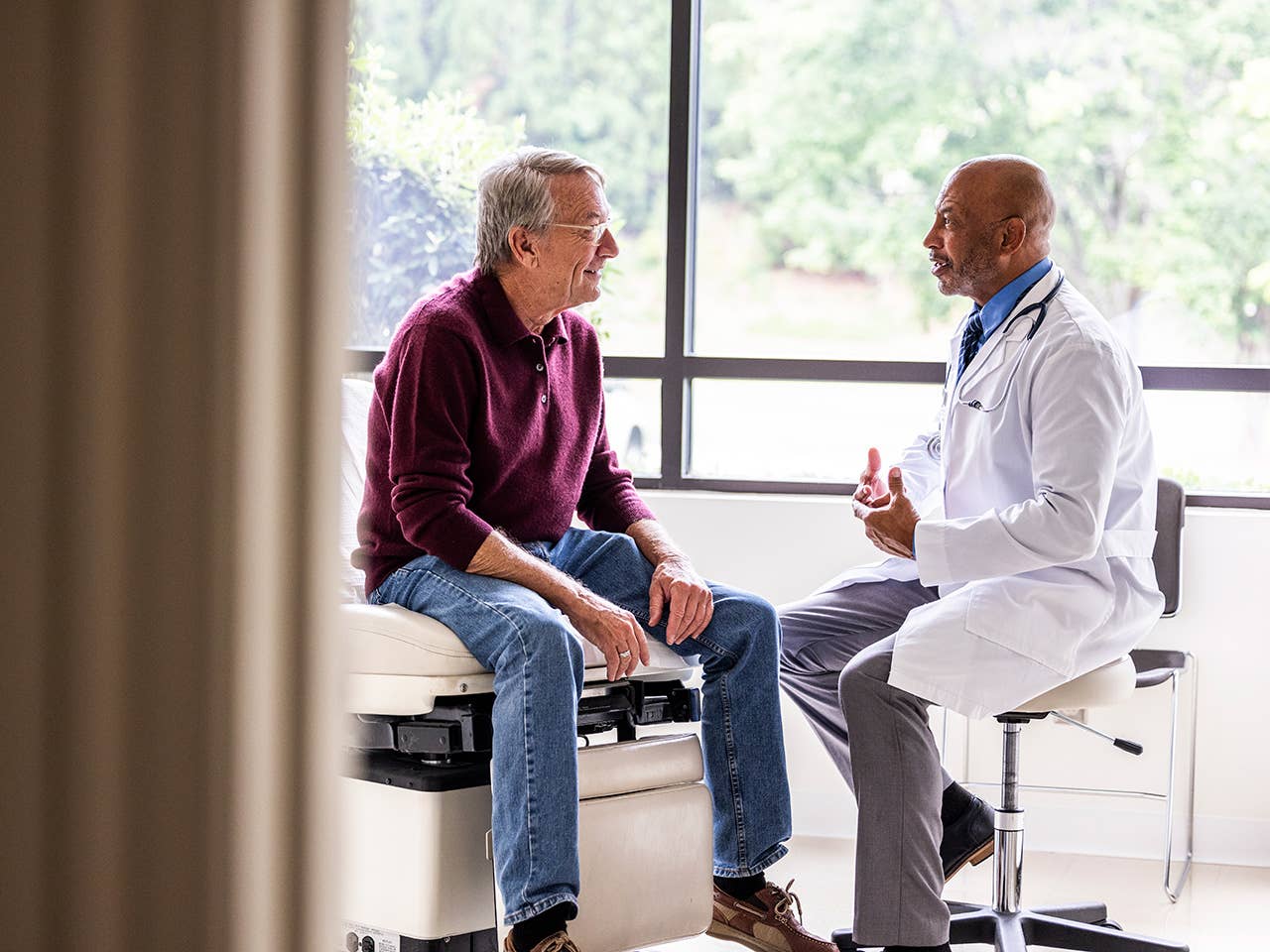 Elderly man wearing glasses and a maroon sweater sits on exam table speaking with male doctor