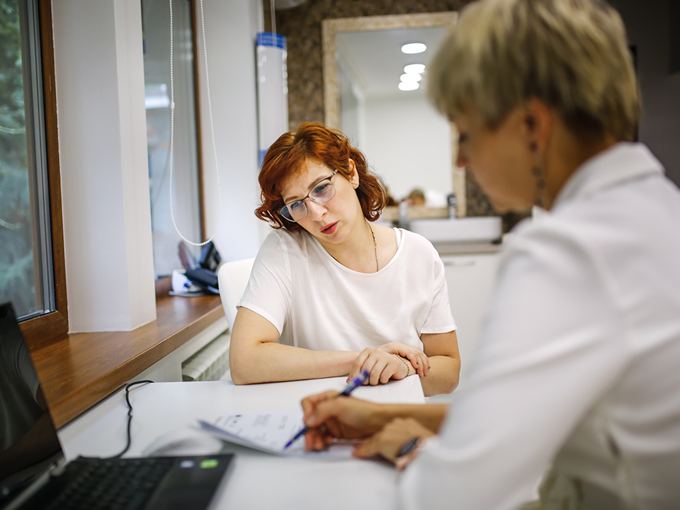 A woman with glasses sitting at a desk, engaged in conversation with a physician.