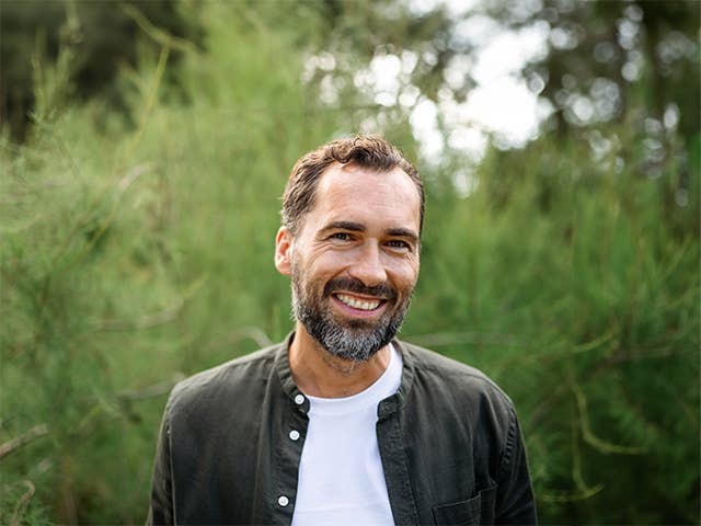 Smiling man with brown hair and a beard standing in outdoors in front of trees