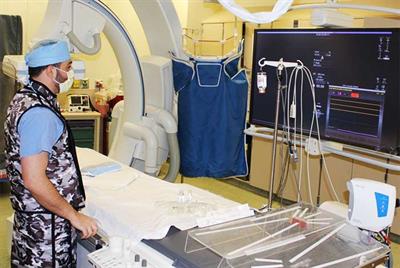 A medical professional in scrubs and a mask stands in a hospital room with diagnostic equipment and a screen displaying data.