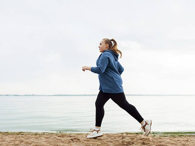 Middle aged white woman in long pants and a sweatshirt jogs alongside a lake
