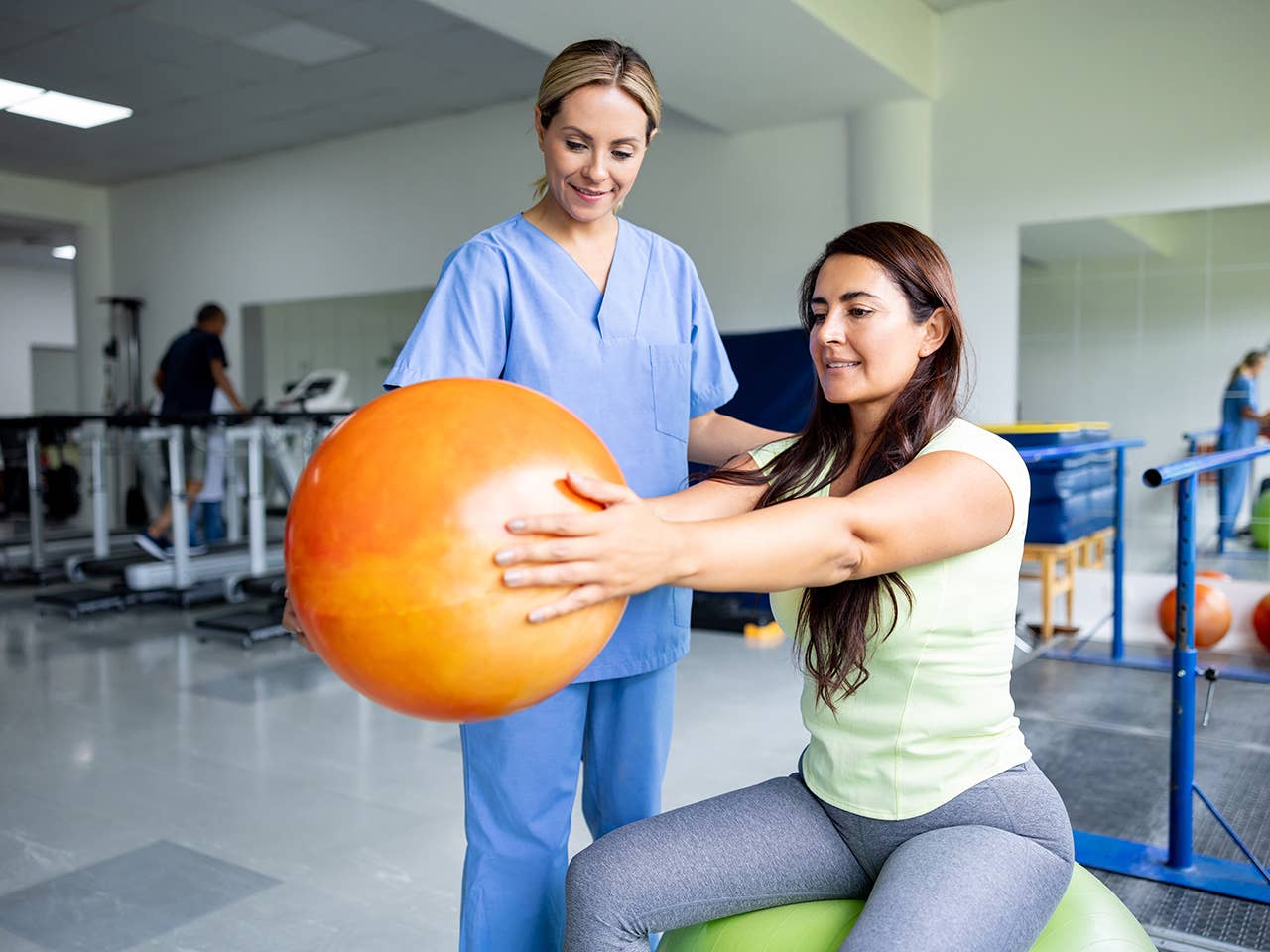 Therapist in light blue scrubs assists a woman holding an inflatable exercise ball in her outstretched arms