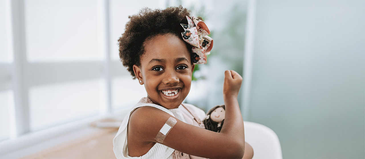 Child showing a bandage after a flu vaccine, holding a doll and flexing an arm in a healthcare setting.