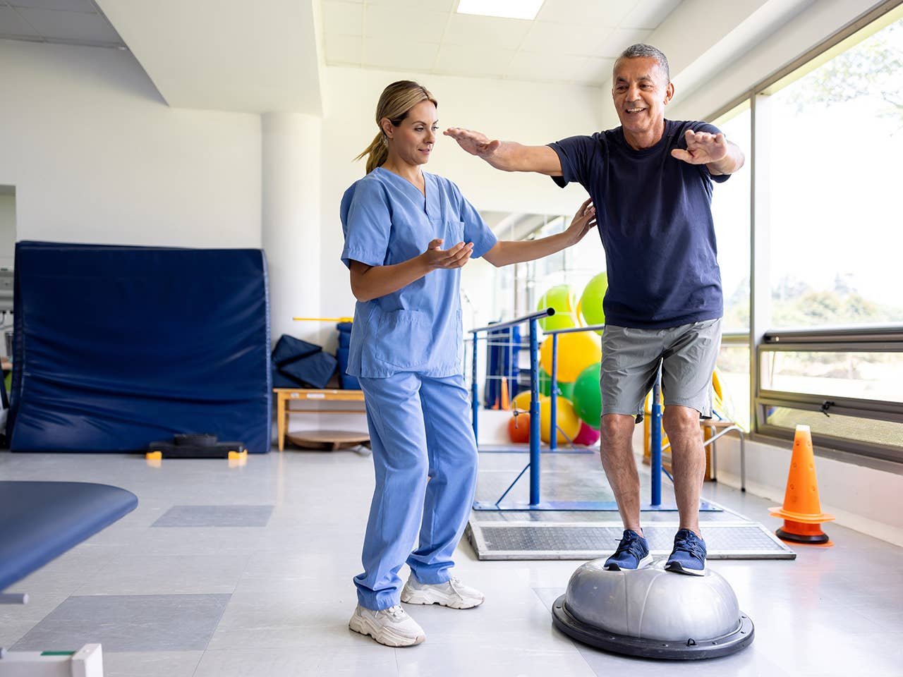 Woman in light blue scrubs helps a man as he balances on equipment in a rehabilitation gym