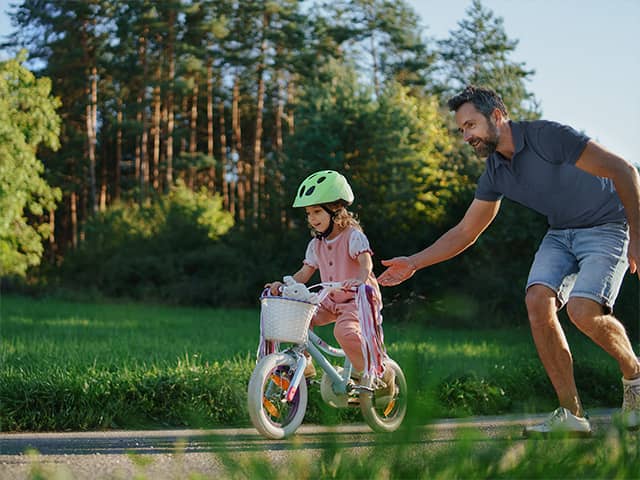 Young white girl wearing a pink outfit and a light green helmet rides a bicycle as her father stands behind her