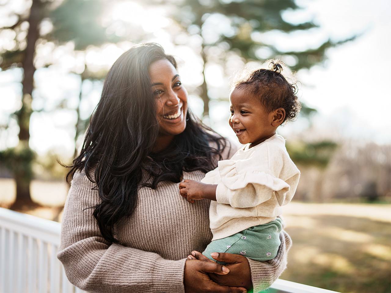 Smiling Black woman wearing a beige sweater holds her daughter as they stand outdoors in the sunshine