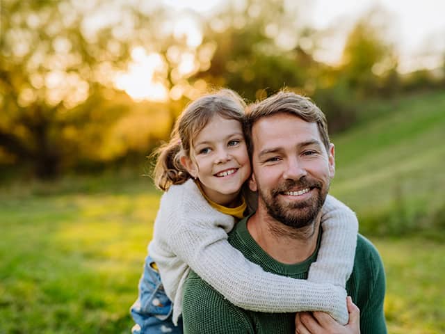 Smiling man with brown hair and a beard carries his smiling daughter on his back while they are outdoors in the sunset