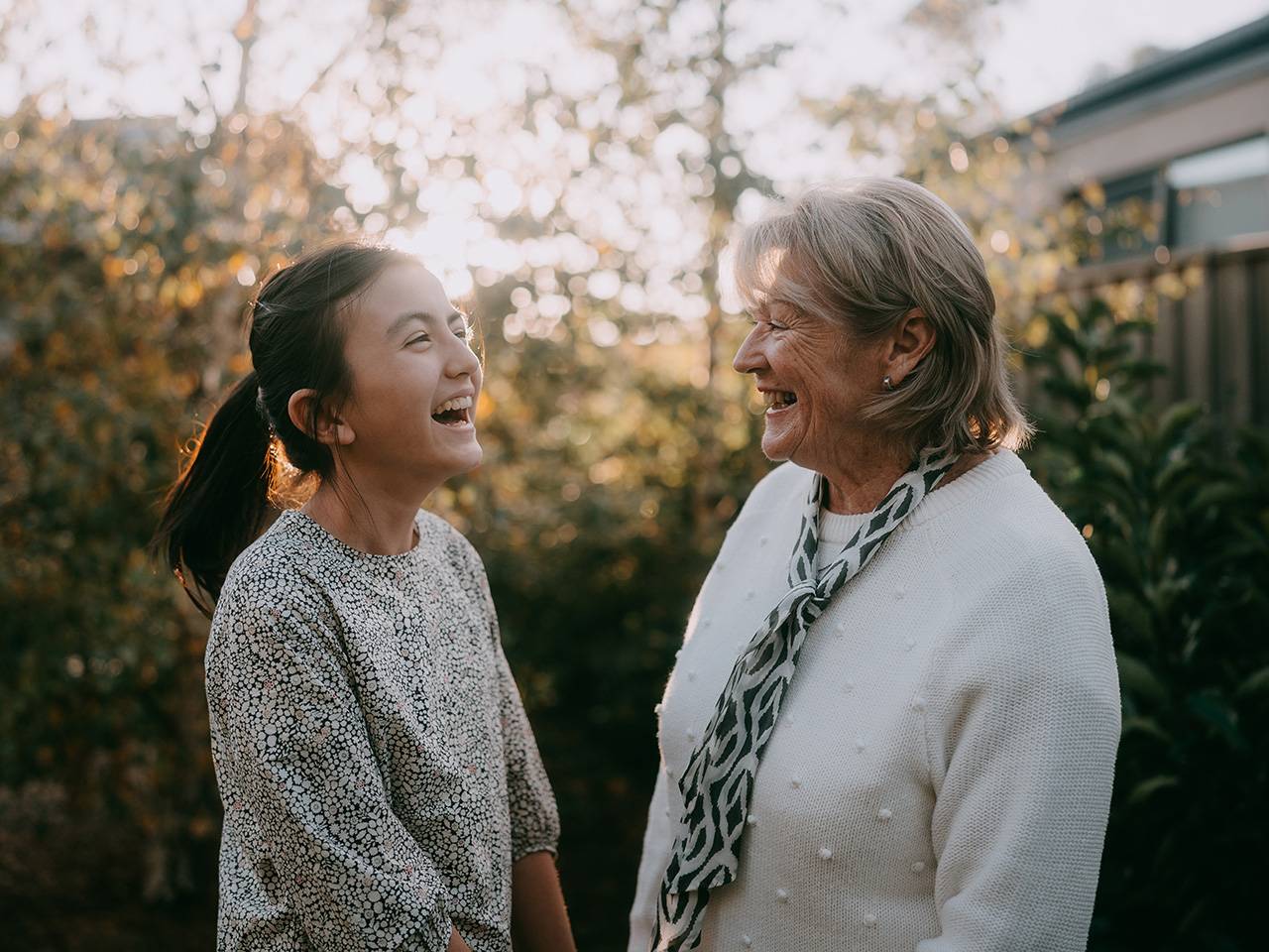 A teenage girl with black hair in a ponytail laughs while speaking to her grandmother outdoors as the sun sets behind trees