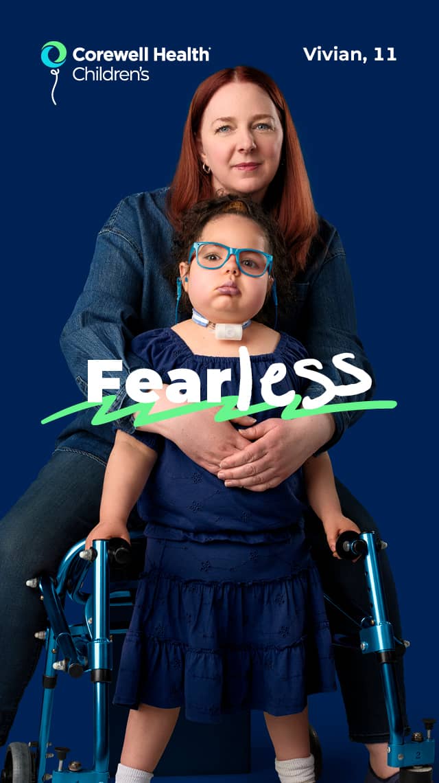 Vivian, an 11 year old patient wearing a blue blouse and light blue glasses, stands with a walker with her mom behind her