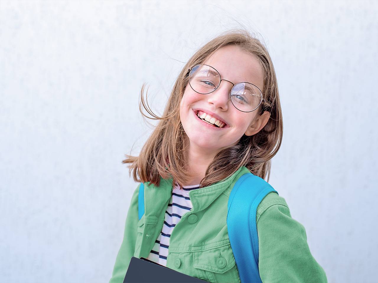 Preteen girl wearing a green shirt and glasses stands in front of a white wall outdoors