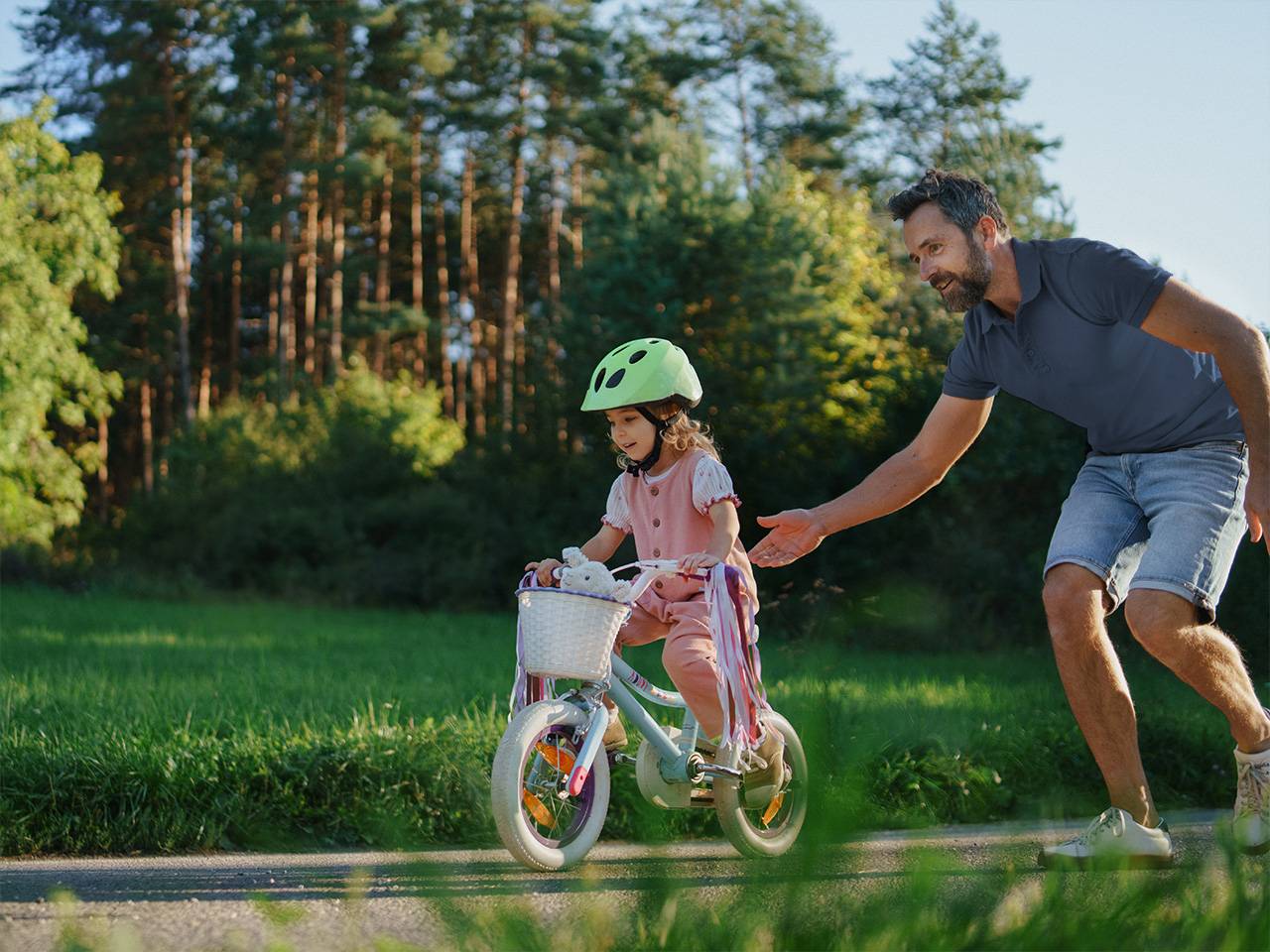 Young white girl wearing a pink outfit and a light green helmet rides a bicycle as her father stands behind her
