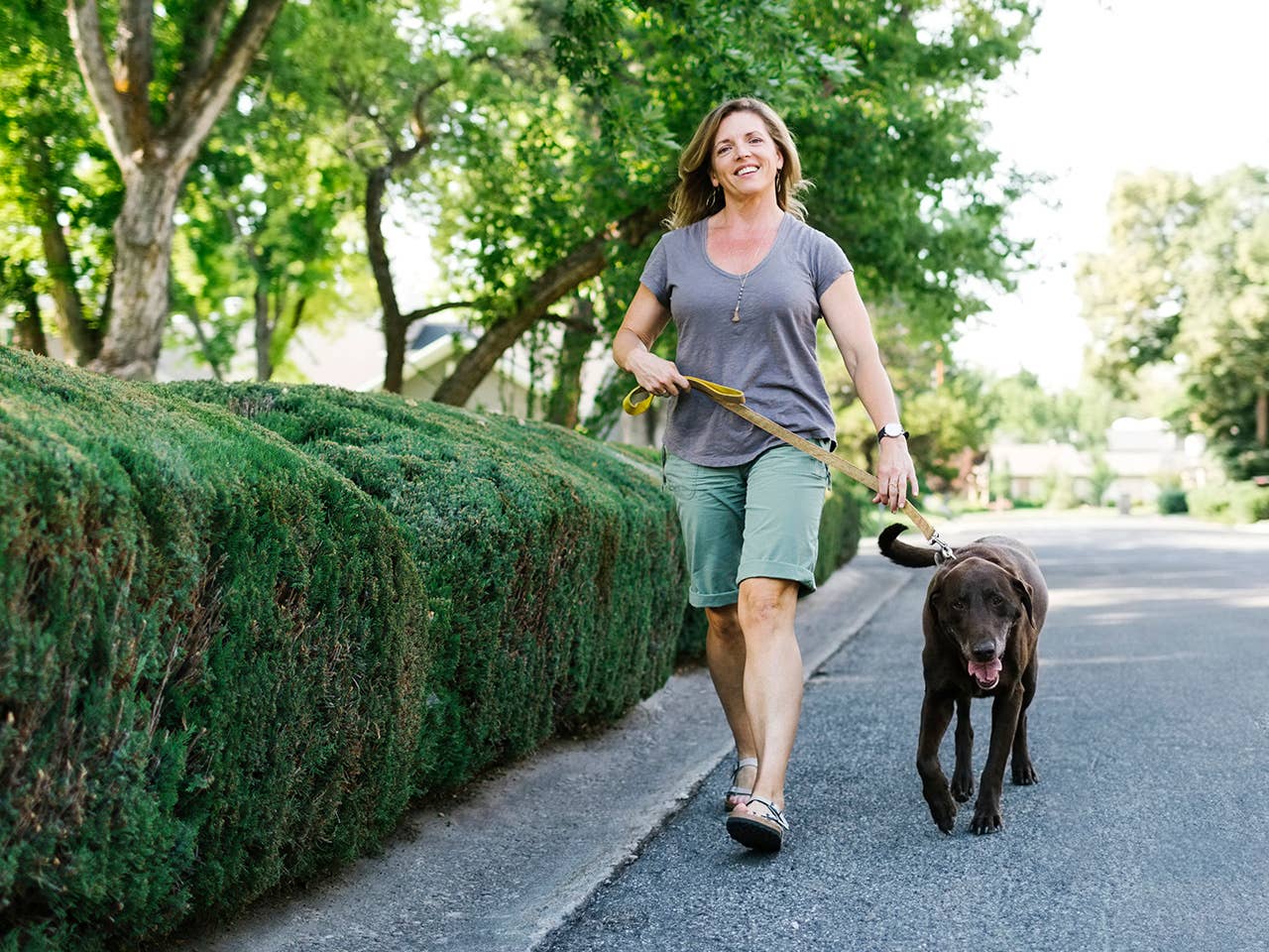 Smiling middle aged white woman walks a black dog in the shade of trees on a sunny day