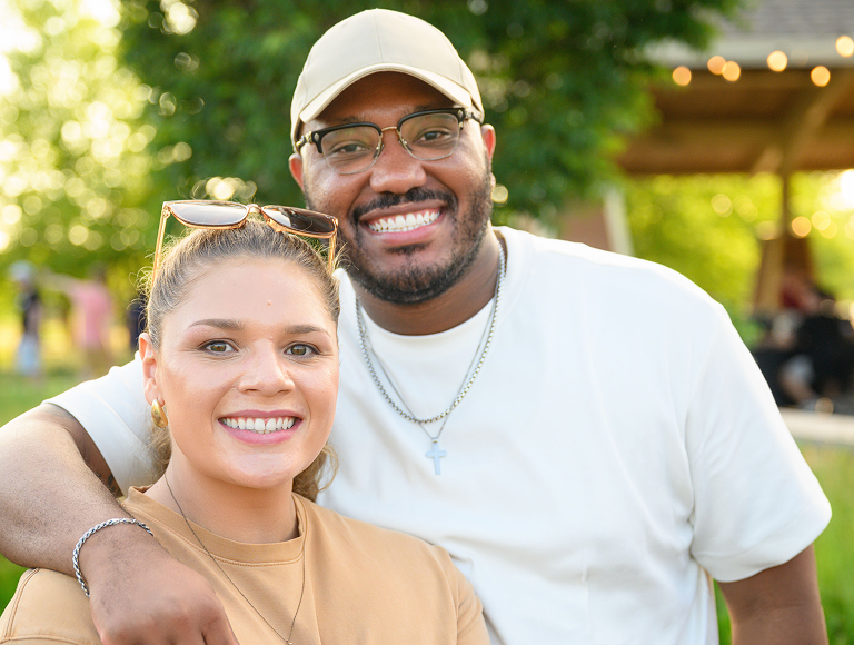 Two individuals standing close together outdoors during a summer event. One person is wearing a tan shirt with a visible name tag and a smartwatch, while the other is wearing a white t-shirt and a necklace with a cross pendant. The background shows green trees and a wooden pavilion with string lights, suggesting a casual outdoor gathering.
