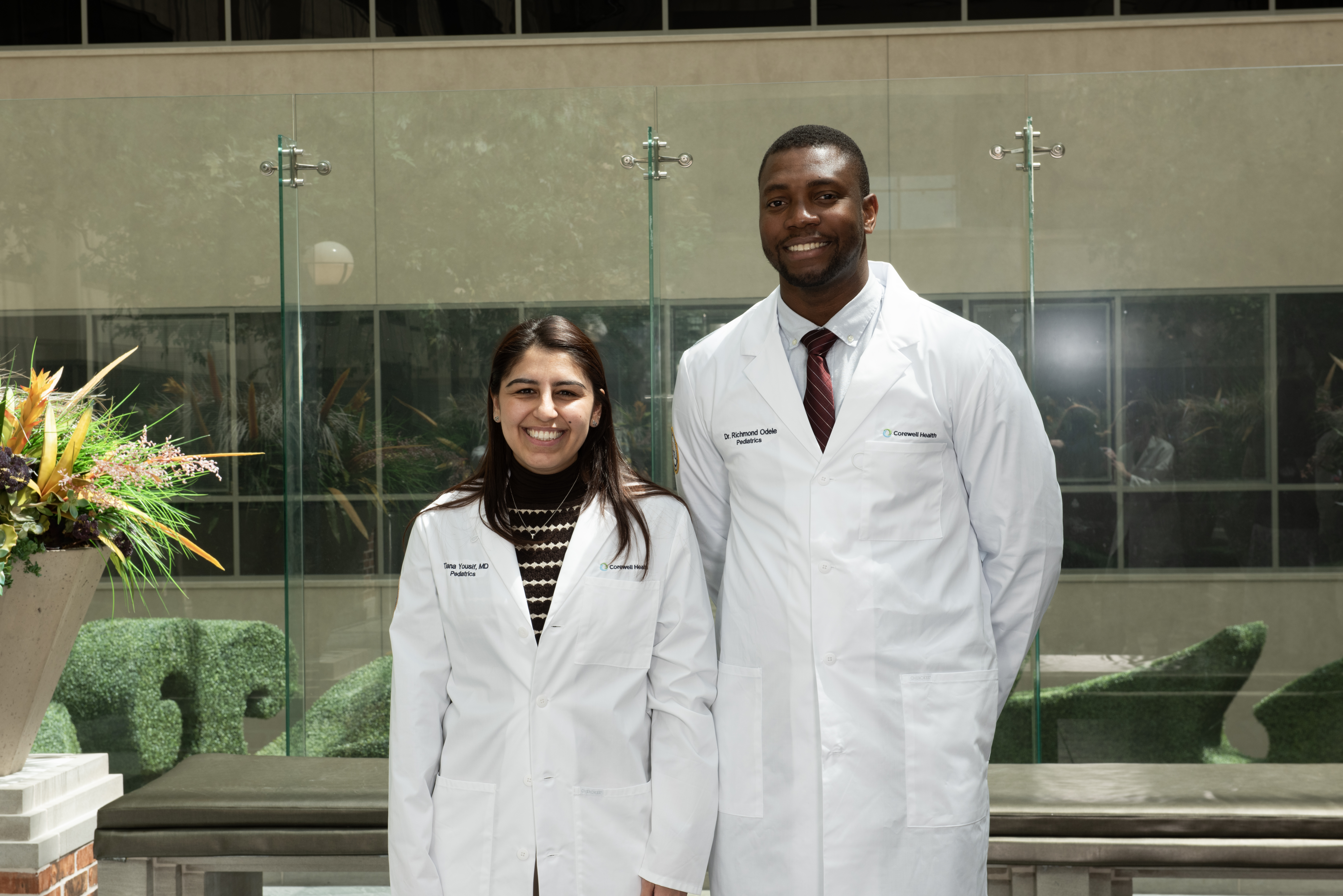 Two pediatric chief residents in white lab coats standing in a hospital atrium with glass walls and indoor landscaping.