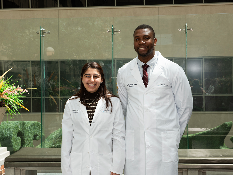 Two pediatric chief residents in white lab coats standing in a hospital atrium with glass walls and indoor landscaping.