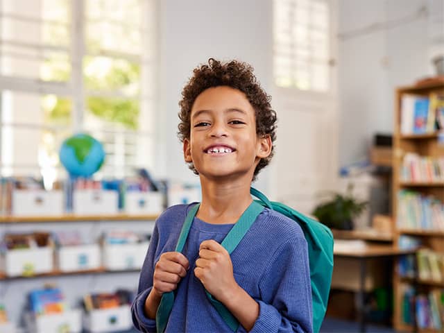 Smiling Black boy wearing a blue shirt stands with his backpack inside a classroom