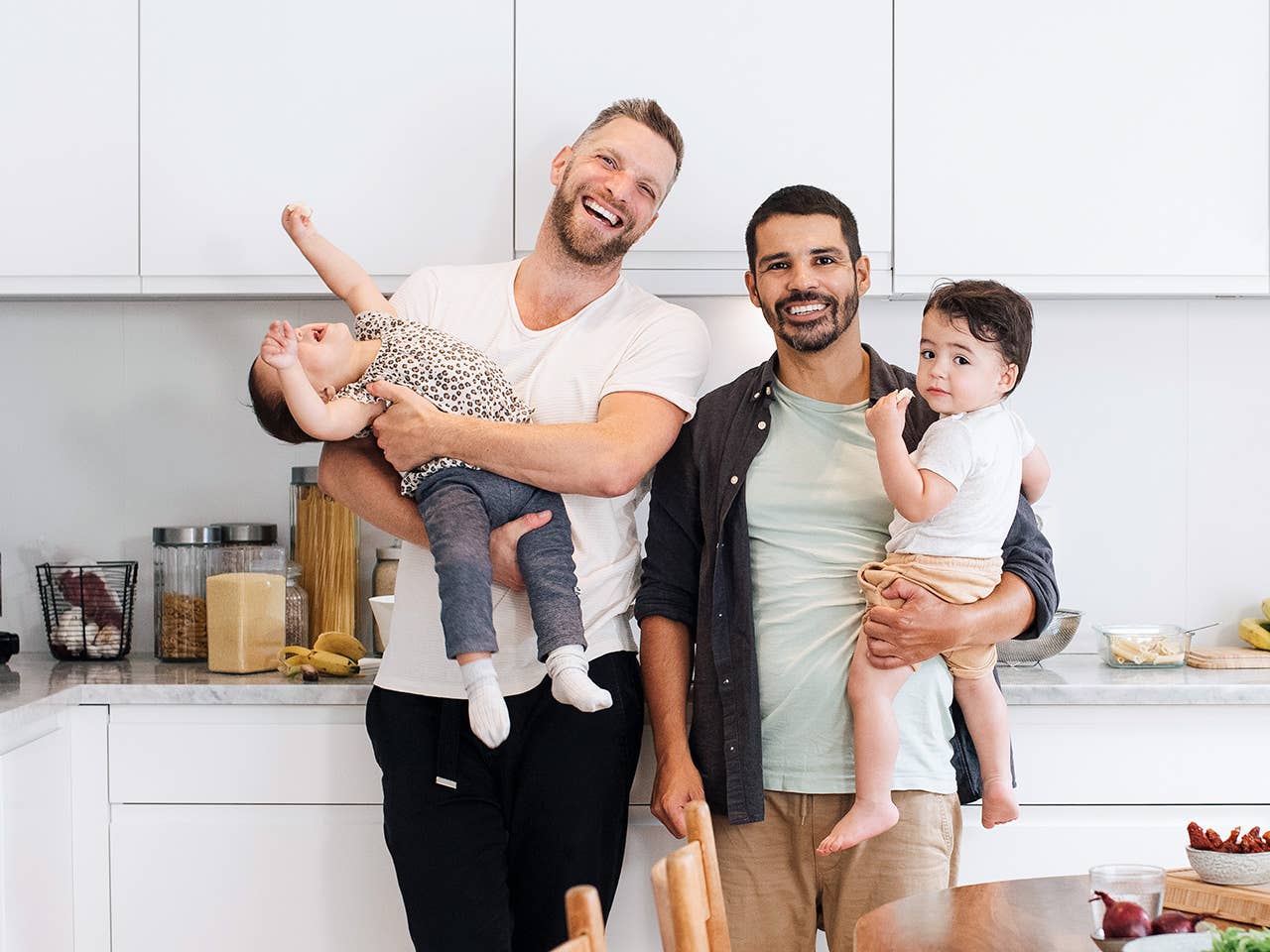 Two smiling men stand side by side as they lift up and hold their young children