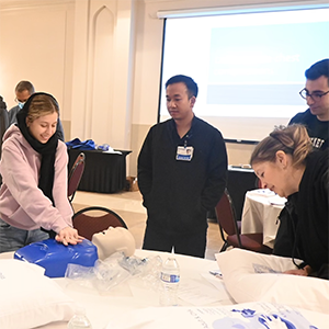 A diverse group of individuals gathers around a table, engaging in a CPR training session with pillows as practice tools.