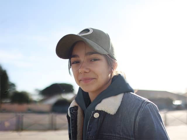 A teenager wearing a denim jacket and a ballcap smiles as they stand outdoors in the sunshine