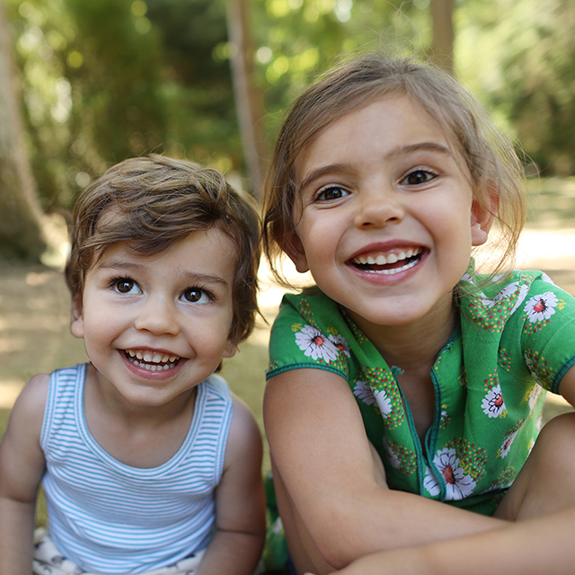 Two children sitting close together outdoors in a wooded park setting, facing the camera.