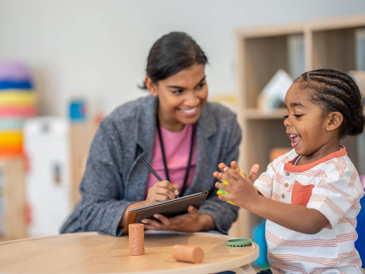 Smiling Black girl claps her hands while seated next to a smiling Corewell Health child life specialist