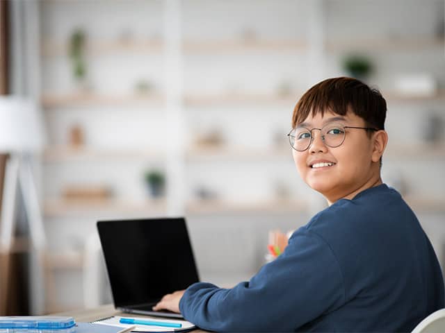 A teenage boy with brown hair and glasses turns and smiles while he sits with a laptop at a school desk