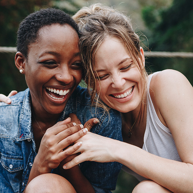 Two young women lean against one another laughing and smiling