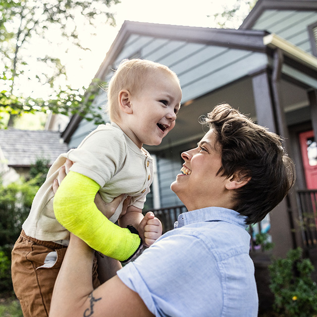 A smiling toddler with a yellow cast on its right arm is lifted up by a smiling woman