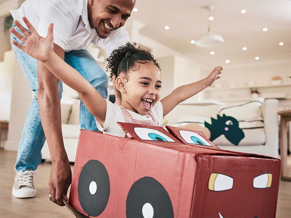 Adult assisting a child playing in a cardboard car indoors.