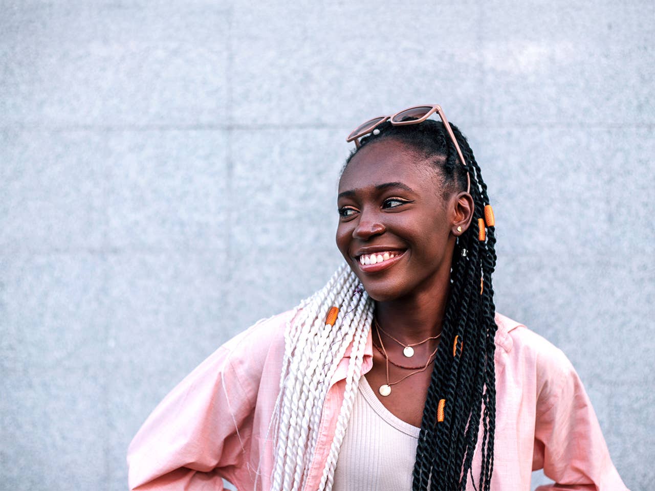 Smiling young Black woman with braided hair stands outdoors in front of a grey wall