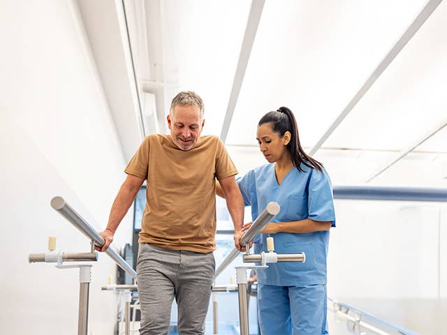 Corewell Health orthopedics rehabilitation team member helps a patient balance with two parallel bars at his side as he stands