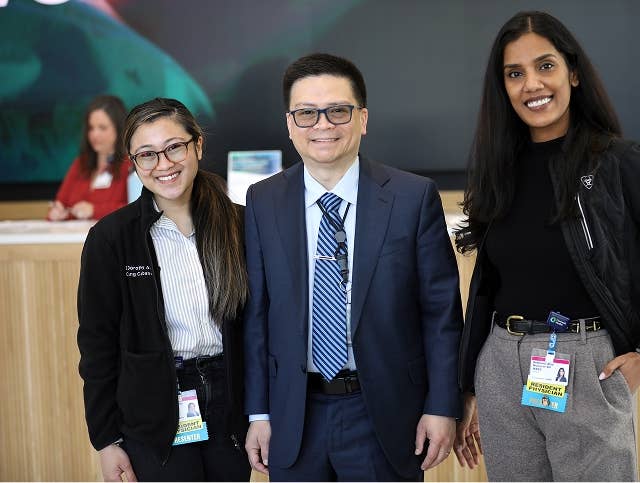 Three Corewell Health team members standing together in a modern lobby area, wearing professional attire and ID badges, with a reception desk and digital display in the background.