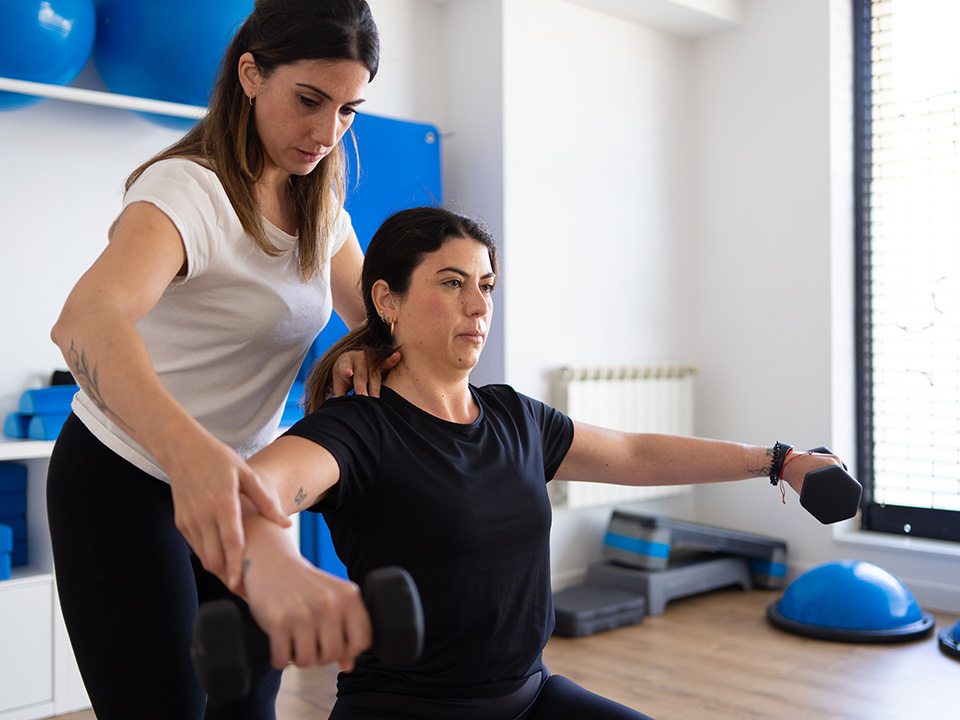 Therapist supports a woman from behind as she lifts hand weights with outstretched arms