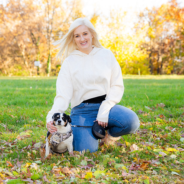 Young white woman kneels down in a grassy field next to a dog