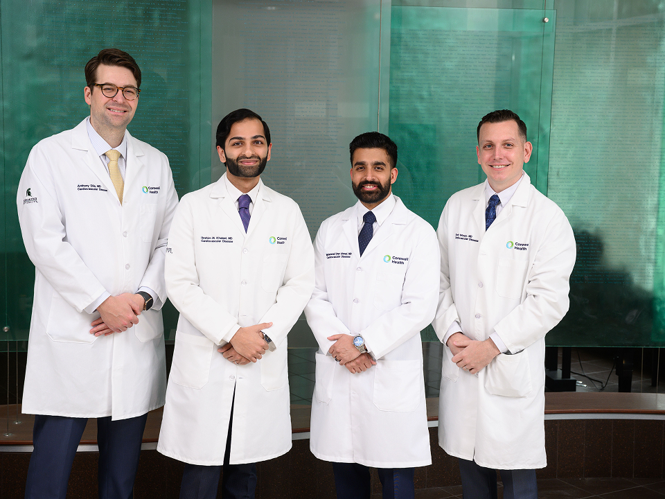 Four medical professionals in white lab coats standing together indoors in front of a glass wall.